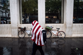 A person wrapped in a red and white striped blanket walks through a concrete colonnade past a parked bicycle.