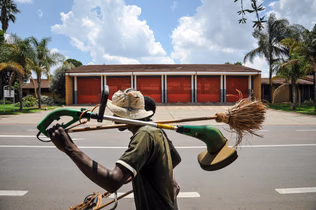 A man walks past a red building carrying a large bundle of grass brooms across his shoulder.