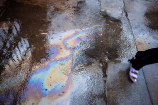 An oil slick spreads bands of colour across a puddle on tar, the edge of a foot at the frame edge.