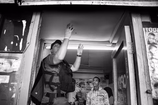 Photograph taken from below of a police officer in a tactical vest with both hands raised in a shop doorway, two men standing inside behind him.