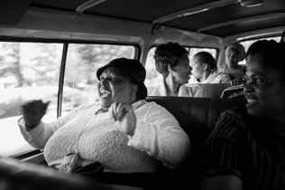 A woman in a cap laughing with her fists raised in the back of a minibus taxi, other passengers talking behind her.
