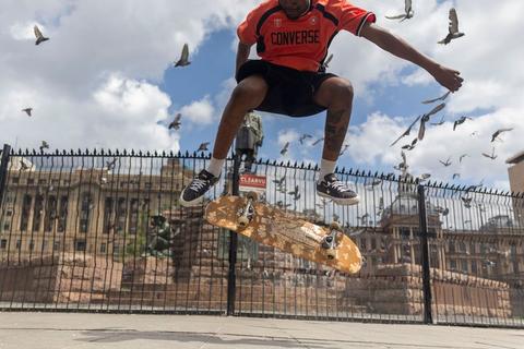 A skater in a red Converse shirt in mid-air above his board, pigeons scattering around him, with a palisade fence and a sandstone government building behind.
