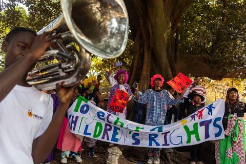 A young brass player with a tuba in the foreground, and behind him people in clown wigs holding a hand-painted banner reading "Together 4 Children's Rights" under a large tree.