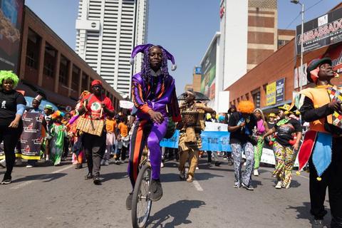 A performer in a purple and orange jester costume rides a unicycle down the middle of a city street, with a costumed parade and banners following behind.