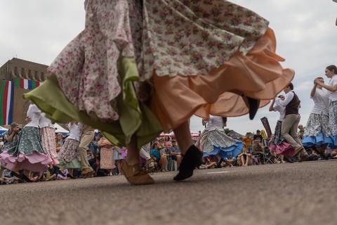 Low-angle view of folk dancers in long floral skirts and boots turning on tarmac, with a seated crowd and festival tents behind.