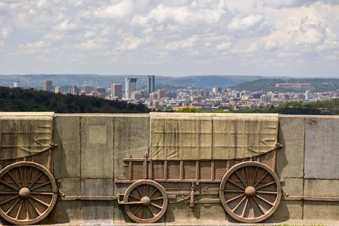 A carved stone relief of ox wagons on the Voortrekker Monument's outer wall, with the Pretoria skyline and hills visible behind it.