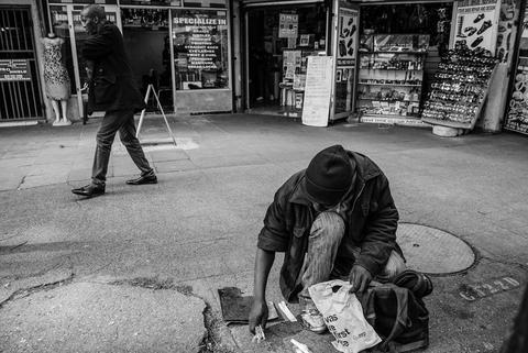 Black and white photograph of a person bent double gathering something from the pavement in front of a wall covered in posters.