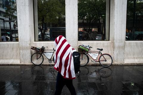 A person wrapped in a red and white striped blanket walks through a concrete colonnade past a parked bicycle.