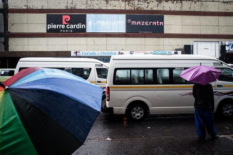 Open umbrellas beside a white minibus taxi in the rain, shopfront signage above.