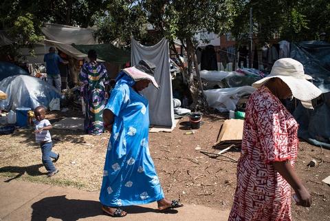 Two women in blue and pink dresses walk along a sand path past corrugated iron shacks.