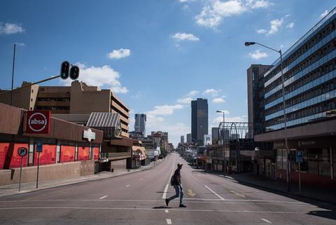 A single person crosses an empty multi-lane street at a traffic light, tall city buildings and blue sky behind.