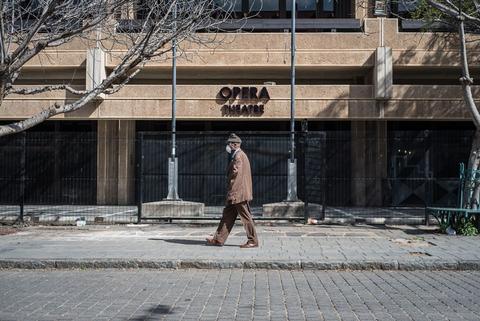 A man walks along an empty pavement past a building signed Opera, one pigeon in the air.