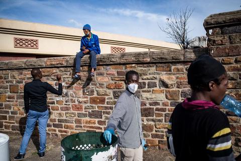 People gathered along a low brick wall, one climbing over it, several wearing face masks, blue sky above.