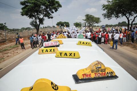 Elevated view over the roofs of yellow minibus taxis, a crowd marching along the road behind them.