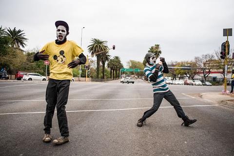Two men move in the middle of an empty street, one in a yellow shirt, trees and buildings behind.