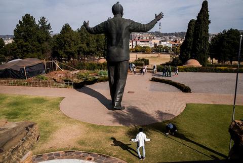 A large bronze statue of Nelson Mandela with arms outstretched on a lawn, a small white dog on the grass in front of it, city visible beyond.