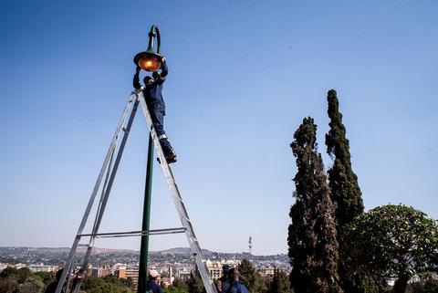 A person works near the top of a metal lighting mast against a clear blue sky.