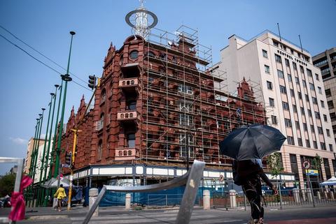 A red brick heritage building wrapped in scaffolding, a person with an umbrella on the pavement below.