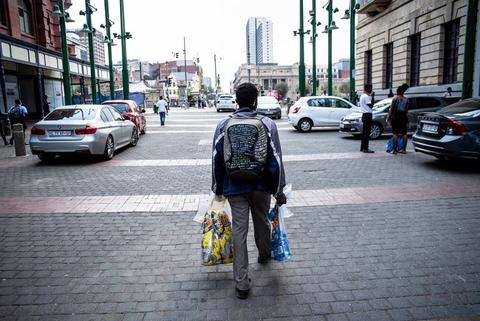 A man walks along a paved street carrying shopping bags, city towers ahead of him.