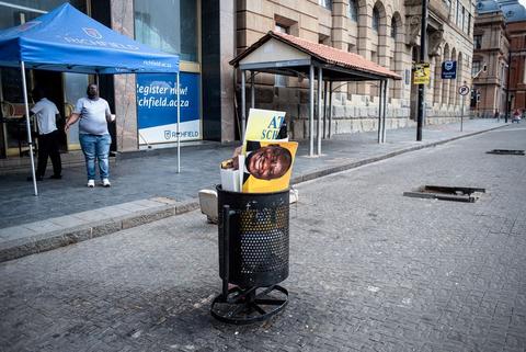 Ramaphosa in the trash bin with a blue gazebo over a street stall on an empty pavement, an older building behind.