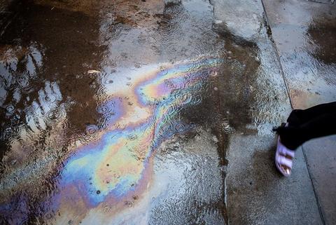An oil slick spreads bands of colour across a puddle on tar, the edge of a foot at the frame edge.