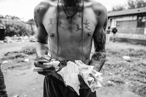 Black and white photograph of a shirtless man with tattooed chest and shoulders, holding a cloth, washing outdoors.