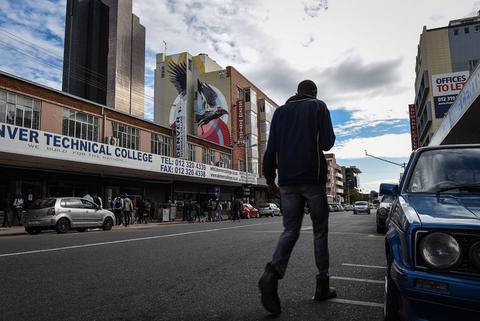 A man walks away down a city street lined with high-rise buildings and painted signage, cars parked at the kerb.
