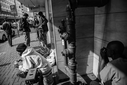 Black and white photograph of a person lying on bedding in a doorway while others stand nearby on the pavement.