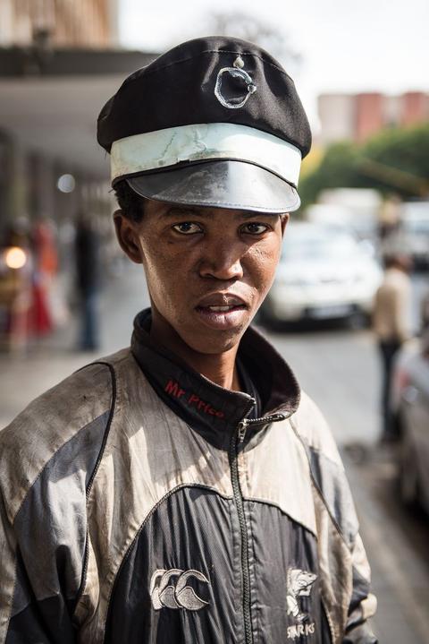 A young man in a peaked cap with a metal badge and a silver jacket looks directly at the camera on a street.