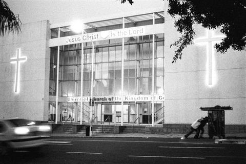 A church facade with two illuminated crosses and a lit sign, a blurred car passing and a man pushing a trolley on the right, Cape Town.