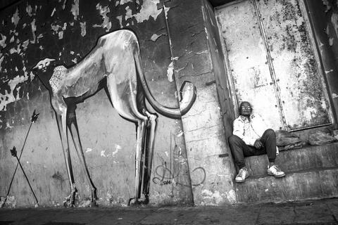 a large painted leopard and arrows on a peeling wall, with a man in a white jacket sitting on a low ledge in the doorway to the right, Woodstock, Cape Town.