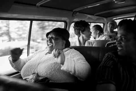 A woman in a cap laughing with her fists raised in the back of a minibus taxi, other passengers talking behind her.