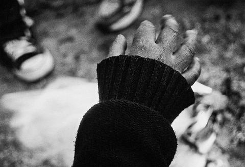 Close-up black and white photograph of a hand in a dark knitted cuff held open above a fire, with other hands out of focus behind.