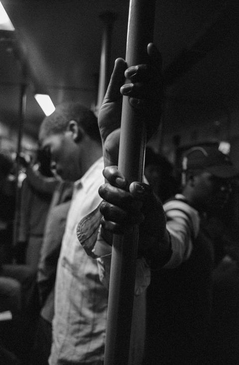 Two hands gripping a vertical metal pole inside a crowded train, passengers out of focus behind.