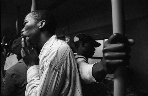 A young man in a striped shirt in profile on a crowded train, one hand to his cheek, another passenger holding the pole beside him.