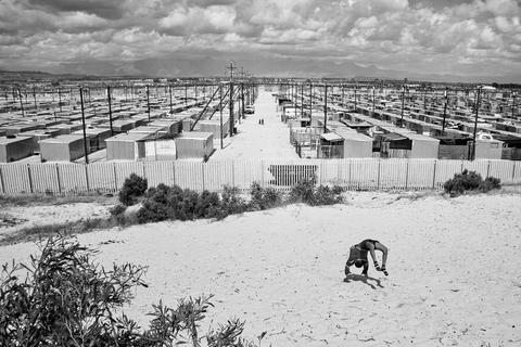 Blikkiesdorp - a large settlement of identical corrugated iron structures in ruled rows, with mountains behind and a boy bending over backwards on sand in the foreground.