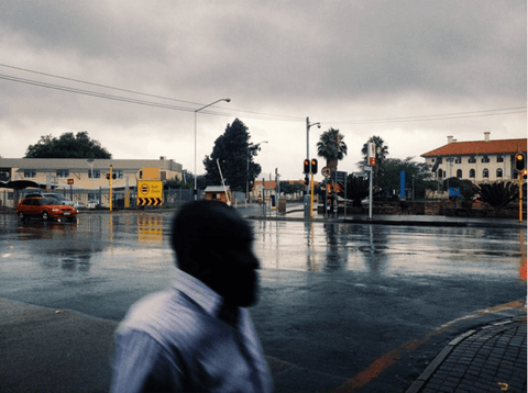 A man in a white shirt walks through a rain-soaked intersection at dusk, his head a dark silhouette against wet tar.