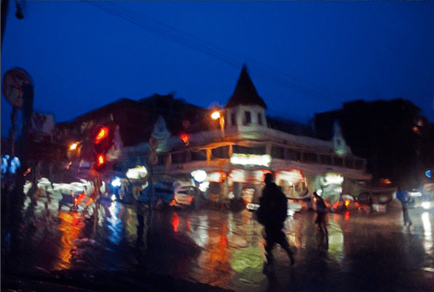 Blurred blue-hour street scene in rain, taxi and shop lights reflecting on a flooded road, a dark figure walking across the centre.