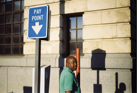 A man in a green golf shirt walks past a blue "Pay Point" sign, with his shadow and the sign's shadow falling on a sandstone wall.