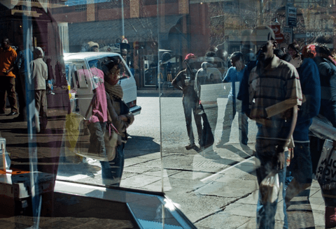 Pedestrians and a minibus taxi seen through and reflected in a shopfront window, layering two views of a busy street.