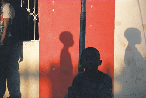 A young boy in silhouette sits against a red and white painted wall, with two head-and-shoulder shadows falling on either side of him.