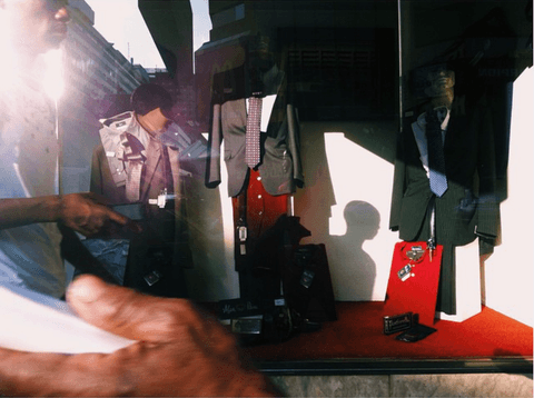 A menswear display window of suits and ties on hangers, a passer-by reflected in the glass and a head-shaped shadow on the back wall.