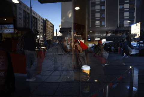 A man sits with a takeaway coffee cup behind a window, the reflected street and buildings overlaying his face and jacket.