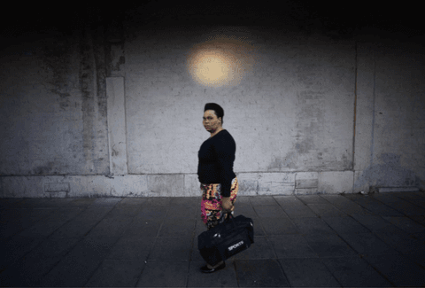 A woman in a black top and patterned skirt walks along a pavement carrying a sports bag, beneath a round pool of light on a dark wall.