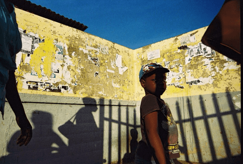 A boy in a cap walks past a yellow wall covered in torn posters, with railing shadows and a figure's shadow cast across the wall.