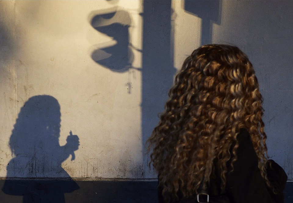 A woman with long curly hair, seen from behind, faces a sunlit wall where her shadow holding a cup falls beside a traffic-light shadow.