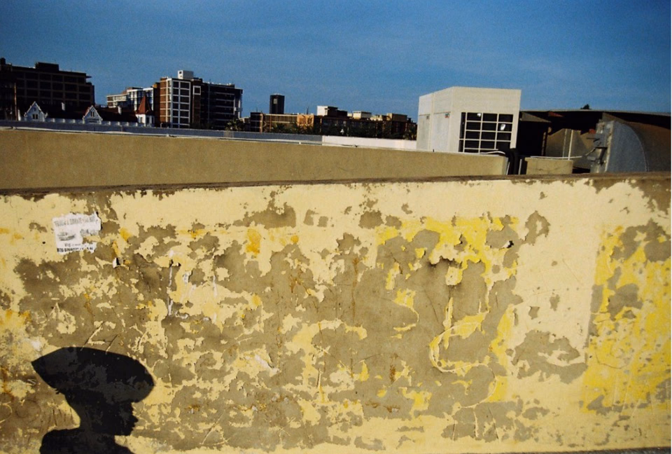 A peeling yellow bridge wall with the Pretoria skyline behind it and the photographer's head-and-shoulder shadow in the lower left corner.