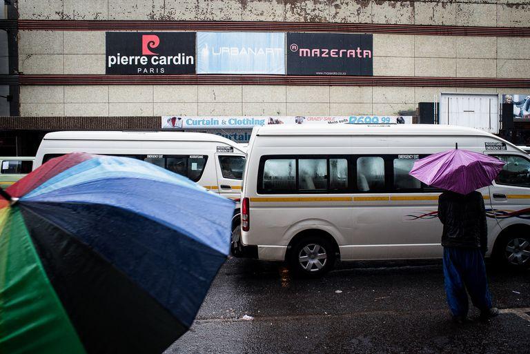 Open umbrellas beside a white minibus taxi in the rain, shopfront signage above.