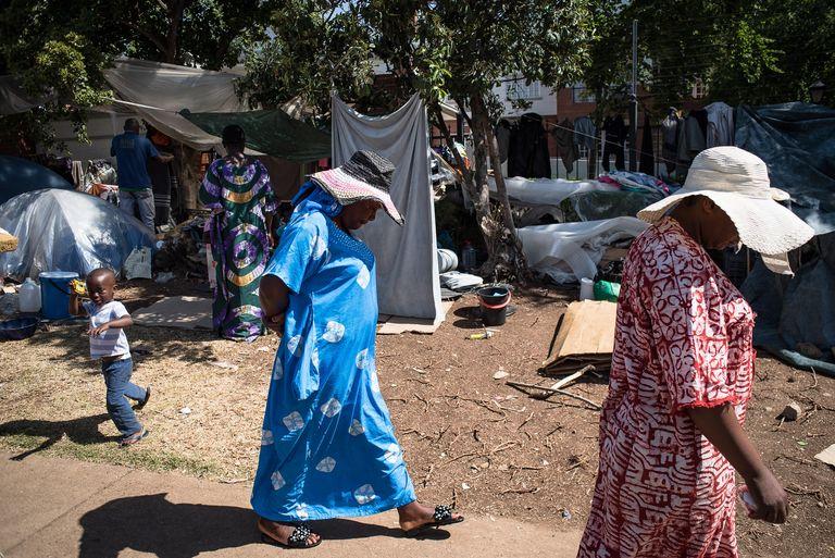 Two women in blue and pink dresses walk along a sand path past corrugated iron shacks.