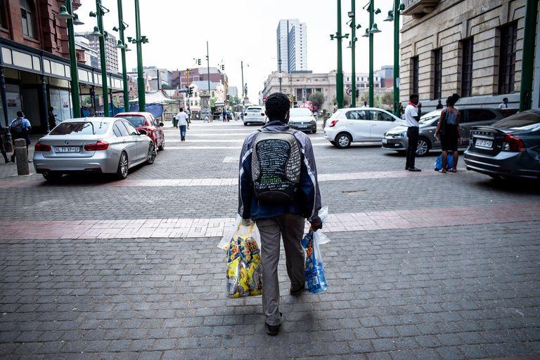 A man walks along a paved street carrying shopping bags, city towers ahead of him.